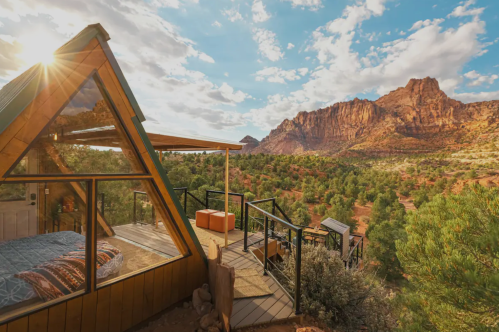 A modern cabin with large windows overlooks a scenic mountain landscape under a bright sky.